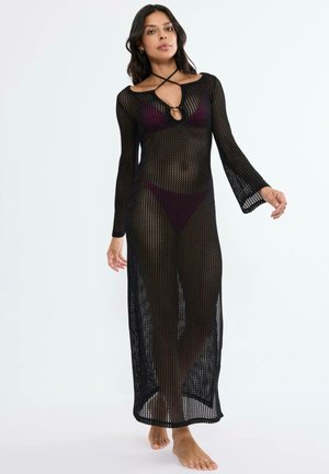 Vrouw draagt een doorschijnende zwarte lange-mouwen gehaakte jurk over een donkerpaarse bikini en poseert blootsvoets tegen een eenvoudige witte achtergrond.