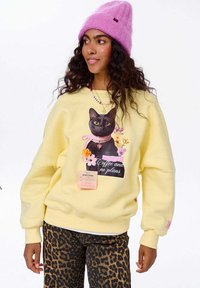 Mladá žena v růžové čepici, žluté mikině s potiskem černého kočky a květin, a kalhotách s leopardím vzorem, hledící do strany.