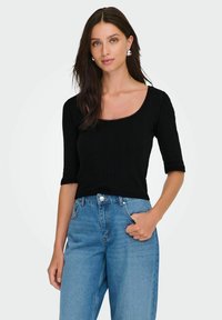 Sort ribbet striktrøje med firkantet halsudskæring og korte ærmer, parret med højttaljede lyseblå denimjeans. Tilbehøret inkluderer sølvøreringe.