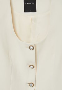 Blusa color crema con scollo rotondo, dotata di chiusura a bottoni con dettagli in perla, realizzata in tessuto morbido e dalla texture liscia.