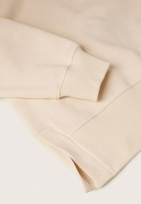 Manche et ourlet inférieur d’un sweat-shirt couleur crème avec poignets côtelés et petite fente latérale sur une surface plane.