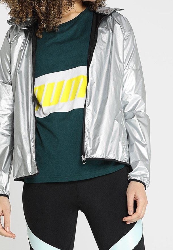 Personne portant une veste argentée brillante par-dessus un t-shirt vert foncé avec un texte jaune vif, associée à des leggings noirs avec des rayures diagonales bleu clair.