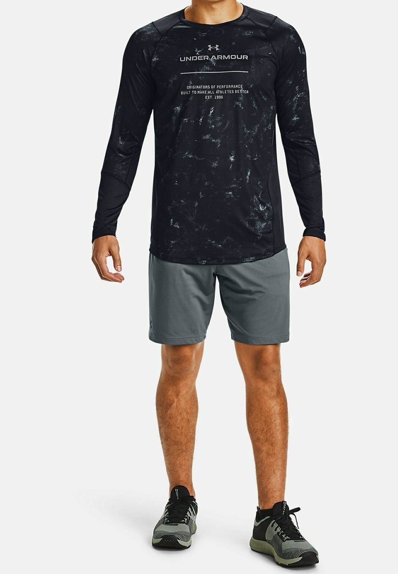 Mężczyzna w czarnej i szarej koszulce z długim rękawem Under Armour, szarych szortach i czarno-szarych butach sportowych stojący na białym tle.