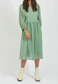 Svetle zelené midi šaty s texturou, balónovými rukávy a naškrobeným pasem. Nosené s bílými kotníkovými botami. Minimalistický design a splývavý silueta.