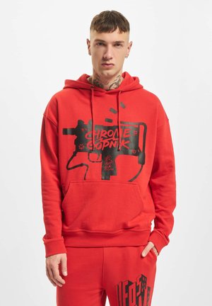 Jeune homme aux cheveux courts et tatouages au cou porte un sweat à capuche rouge et un pantalon de survêtement avec un texte graphique noir et un design, debout sur fond blanc.