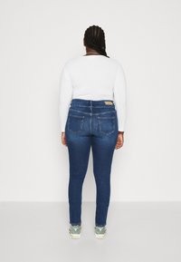 Mujer con cabello trenzado que lleva una camiseta blanca de manga larga, jeans ajustados azules y zapatillas verdes, de pie frente a una pared blanca.