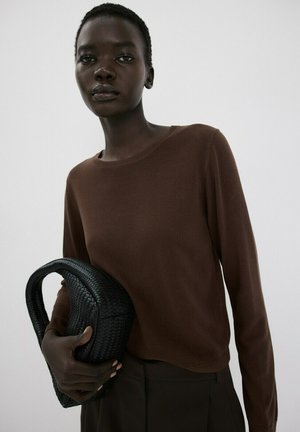 Persona con una blusa marrón de manga larga sosteniendo un bolso tejido negro contra un fondo liso de color gris claro.