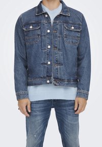 Tummansininen farkkutakki, jossa on kaksi rintataskua, hopeiset napit ja lyhyt muotoilu. Pukeutunut vaaleansinisen villapuseron päälle ja yhdistetty farkkuihin.