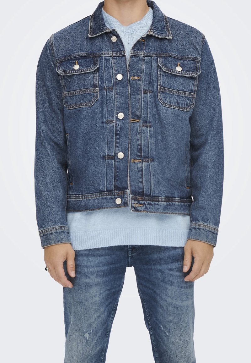 Tummansininen farkkutakki, jossa on kaksi rintataskua, hopeiset napit ja lyhyt muotoilu. Pukeutunut vaaleansinisen villapuseron päälle ja yhdistetty farkkuihin.