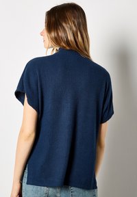 Marineblauwe gebreide top met korte mouwen en een geribde textuur. Beschikt over een hoge nek en een losse pasvorm, met een iets langere achterkant.