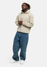 Beige Kapuzenpullover mit langen Ärmeln, kombiniert mit lockeren blauen Jeans und weißen Sneakers. Schlichtes Design und lässige Passform.