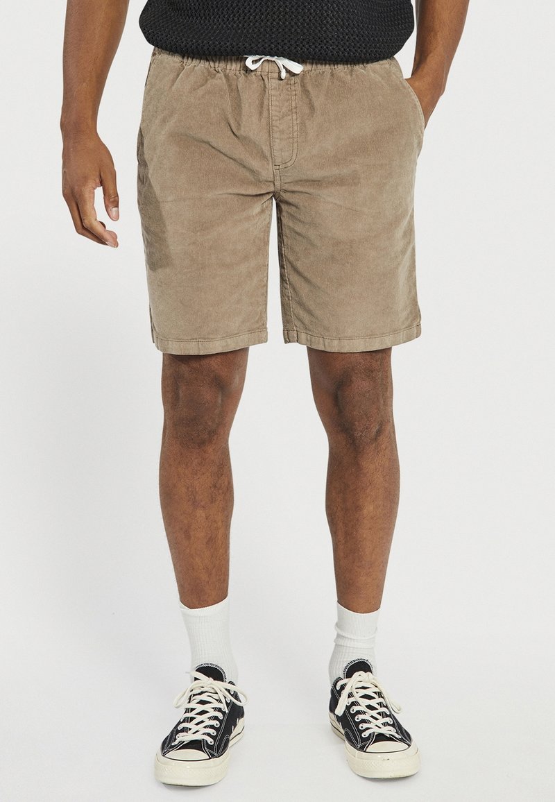 Shorts en velours côtelé marron avec une taille élastique et un cordon de serrage, dotés de poches latérales et d'une coupe décontractée, assortis à des baskets noires et des chaussettes blanches.