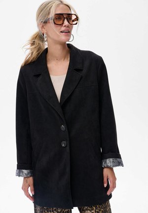 Veste en daim noire avec col cranté, deux boutons de fermeture, poches sur les côtés et doublure des poignets à motif gris. Portée sur un haut clair et un pantalon à imprimé animal.
