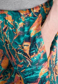 Costume de baie multicolore cu un model abstract vibrant în nuanțe de turcoaz, portocaliu și negru, având talie elastică și buzunare laterale.