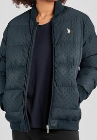 Marinblå pufferjacka med diamantrutigt mönster, ribbad krage, dragkedjestängning och liten logotyp på bröstet. Har quiltade sektioner för isolering.