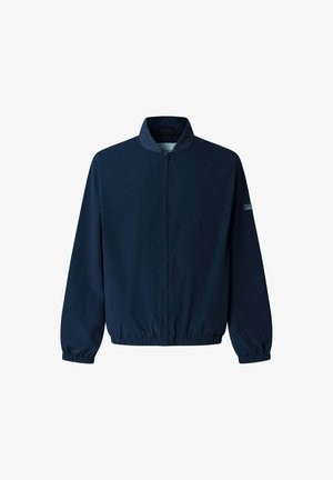Jonge man met een marineblauw bomberjack, lichtblauw gestreept hemd, blauwe jeans en beige casual schoenen, staand tegen een effen lichte achtergrond.