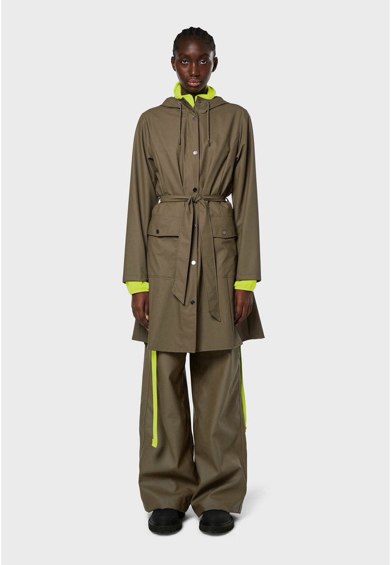 Cappotto impermeabile verde oliva con cappuccio, cintura e due tasche frontali. Presenta accenti giallo neon sulle maniche e pantaloni larghi coordinati.