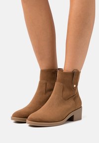 Braune Wildleder-Stiefeletten mit runder Zehenform, niedrigem Blockabsatz, seitlichem Reißverschluss und dekorativer Nahtakzentuierung.