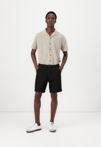 Kortärmad, beige skjorta med ett zickzackmönster, parat med svarta shorts. Vita sneakers med grå accenter fulländar outfiten.