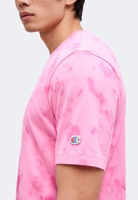 Personne portant un t-shirt manches courtes tie-dye rose avec un petit logo Champion brodé sur la manche gauche, vu de côté.