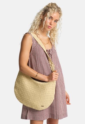 Jeune femme aux cheveux blonds bouclés portant une robe à carreaux rouges et des bijoux en or, portant un grand sac à bandoulière beige tissé.