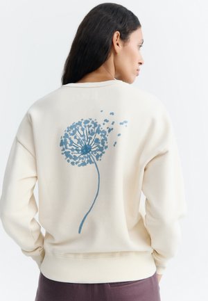 Vrouw met lang donker haar die een crèmekleurige sweater draagt met een blauw paardebloemontwerp op de rug.