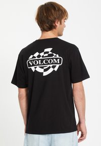 Schwarzes Baumwoll-T-Shirt mit kurzen Ärmeln. Großes weißes "VOLCOM"-Logo mit einem karierten Muster auf dem Rücken. Einfaches, lässig geschnittenes Design.