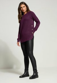 Lila, langärmliger Pullover mit asymmetrischem Saum und drei dekorativen Knöpfen an der Seite, kombiniert mit schwarzen Leggings aus Kunstleder und Ankle Boots.