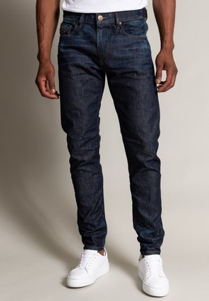 Homme portant un jean slim bleu foncé et des baskets basses blanches, debout devant un fond uni.