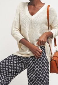 Krémový pletený svetr s výstřihem do V a tříčtvrtečními rukávy, kombinovaný s tmavě modrými vzorovanými kalhotami. Doplňkem je hnědá taška s texturou.