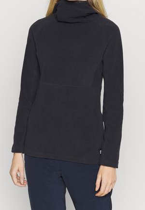 Woman wearing a black long-sleeve turtleneck sweater and dark pants, standing against a plain light background.