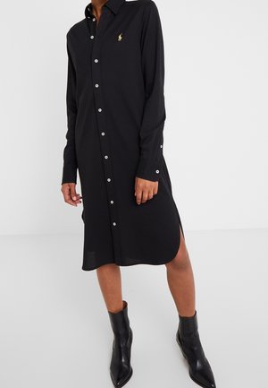 Personne portant une robe chemise midi noire boutonnée, manches longues, fentes latérales et bottines noires.