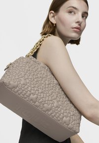 Beżowa torebka z wytłaczanym wzorem misia, o skórzanej fakturze i wyraźnym złotym paskiem na ramię. Posiada wytłoczone logo marki na dnie.