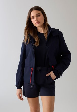 Mujer con cabello largo y castaño, lleva una chaqueta azul marino con tiradores de cremallera rojos, top negro y pantalones cortos azul marino, de pie frente a un fondo claro y sencillo.