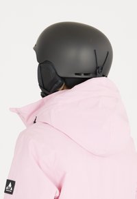 Pink ski jacket with a hood, featuring a black collar and logo patch. A matte black helmet is worn, highlighting ear protection details.