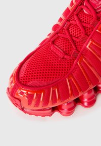 Rdeča športna čevlja z mrežasto zgornjico, ojačanim prstom, edinstvenim razdeljenim podplatom in podrobnim vezanjem na beli podlagi.
