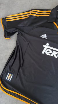Crna majica s narančastim detaljima, s tri pruge na rukavima. Sadrži logo "Teko" i Adidas brand. Lagani materijal.