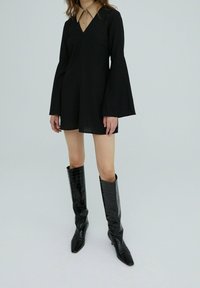 Robe mini noire à manches en cloche avec un décolleté en V profond, superposée à une chemise col beige. Associée à des bottes noires brillantes et texturées de grande taille.
