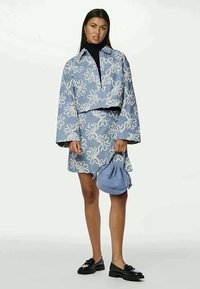 Casaco cropped azul claro e saia combinando com bordados florais brancos. Acompanhados por uma bolsa de tecido azul e sapatos pretos. Textura suave.