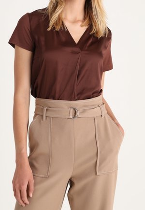 Femme portant un haut marron à manches courtes et col en V, rentré dans un pantalon beige taille haute avec une ceinture assortie et deux poches plaquées à l'avant.