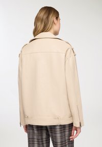 Giacca in pelle beige con ampio colletto, polsini con cerniera e vestibilità comoda. Texture liscia con dettagli di cucitura e fibbie laterali.