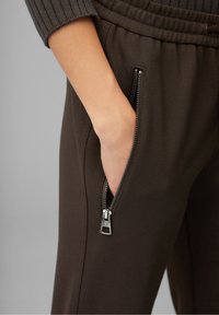 Braune schmal zulaufende Hose aus strukturiertem Stoff, ausgestattet mit Reißverschluss-Seitentaschen und einem elastischen Bund für zusätzlichen Komfort.