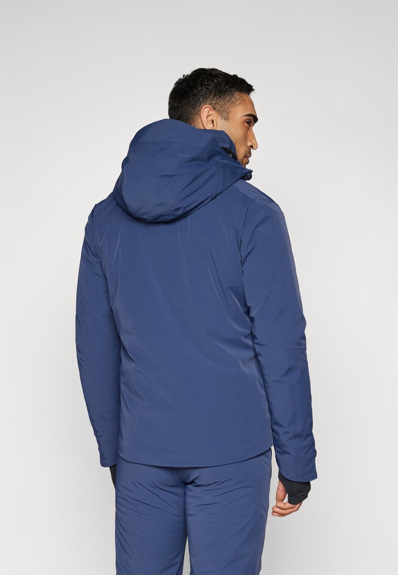 Veste imperméable bleu marine avec capuche, au texture lisse, à design ajusté et aux poignets élastiques contrastants. La vue arrière montre un detailing minimal.