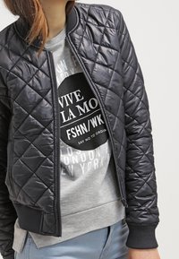 Chaqueta bomber negra acolchada con puños y dobladillo acanalados, que presenta una cremallera frontal. Se lleva sobre una sudadera gris claro con gráficos de texto.