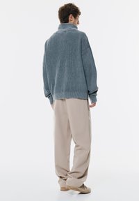 Pull gris oversized côtelé avec un col montant et des manches longues, associé à un pantalon beige clair à coupe décontractée et des chaussures beiges.