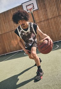 Jugador de baloncesto vistiendo una camiseta sin mangas de patrón blanco y negro y pantalones cortos negros, botando un balón de baloncesto en una cancha con paredes de madera.