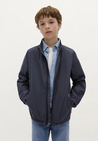 Marineblauwe lichtgewicht jas met een rits aan de voorkant, elastische manchetten, zijzakken en een ontspannen pasvorm, gedragen over een lichtblauw shirt en een witte T-shirt.