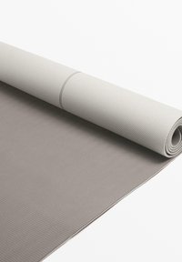 Joga mat v svetlo sivi barvi z teksturirano površino; na enem koncu je zvit, kar razkriva gladko, temneje sivo plast pod njim.