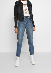 Svart denimjacka, vit t-shirt med grafisk design och matillustrationer, högmidjade blåjeans, vita sneakers och uppvikta ärmar.