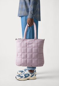 Vadderad rosa tygväska med två robusta handtag, med en strukturerad yta och en logodetalj i mitten. Bärs med jeans och sneakers.